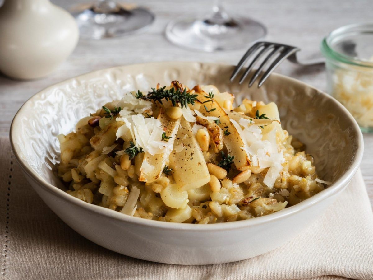 Ein heller Teller mit Gerstenrisotto und Spargelstangen auf einem beigen Untergrund. Dazu ein Glas mit Parmesan und helles Geschirr im Hintergrund.