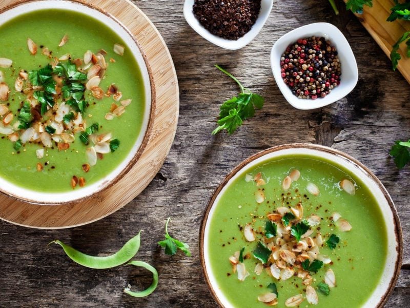 Zwei Teller gefüllt mit einer grünen Gemüsesuppe und dekoriert mit gerösteten Mandeln und Kräutern.