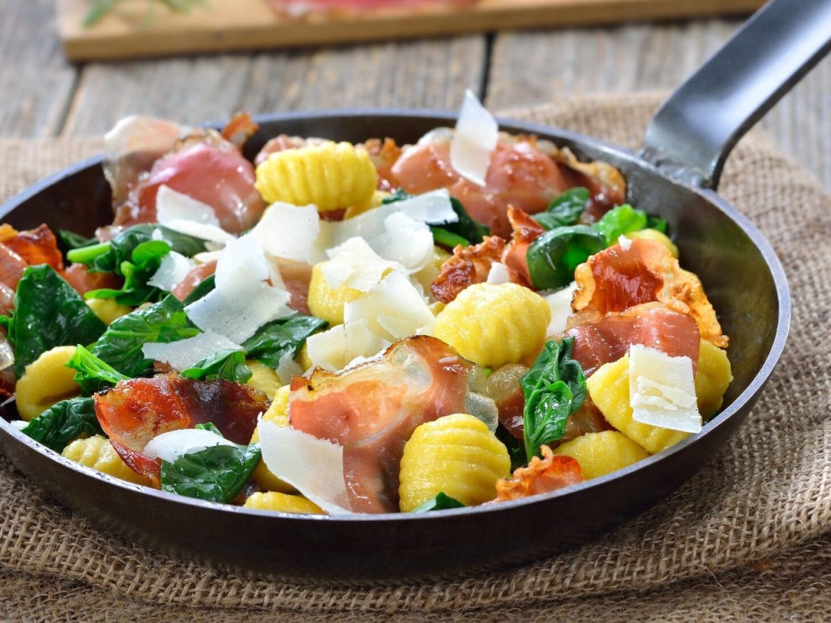 Gnocchi, Spinat, Speck und Parmesan in einer gusseisernen Pfanne. Diese steht auf einem Leinentuch.