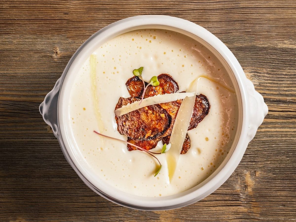 Knoblauchcremesuppe mit gebratener Salami in weißer Schüssel, angerichtet auf Holzplatte.