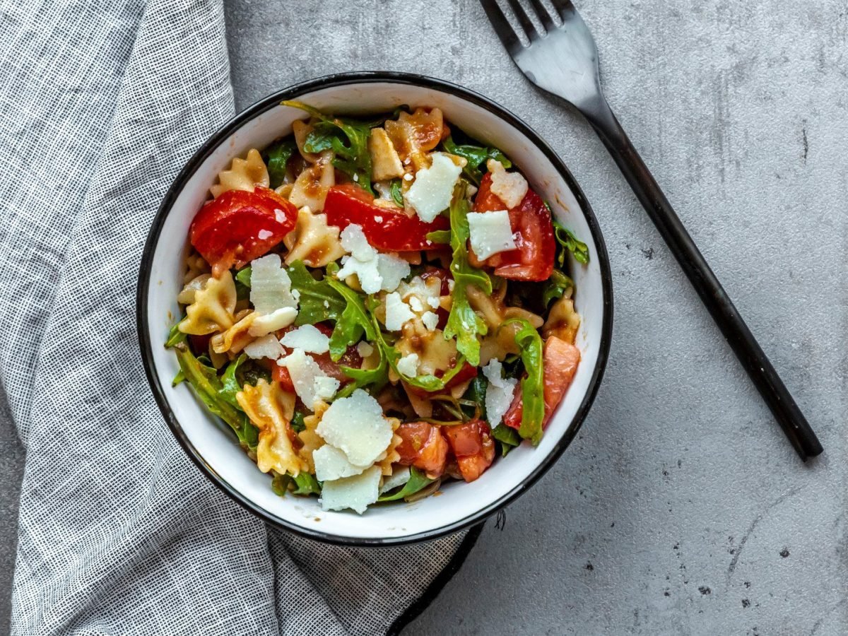 Schüssel mit Nudelsalat, Rucola, Tomate und Parmesan auf grauem Untergrund. Neben liegen ein Tuch und eine Gabel.