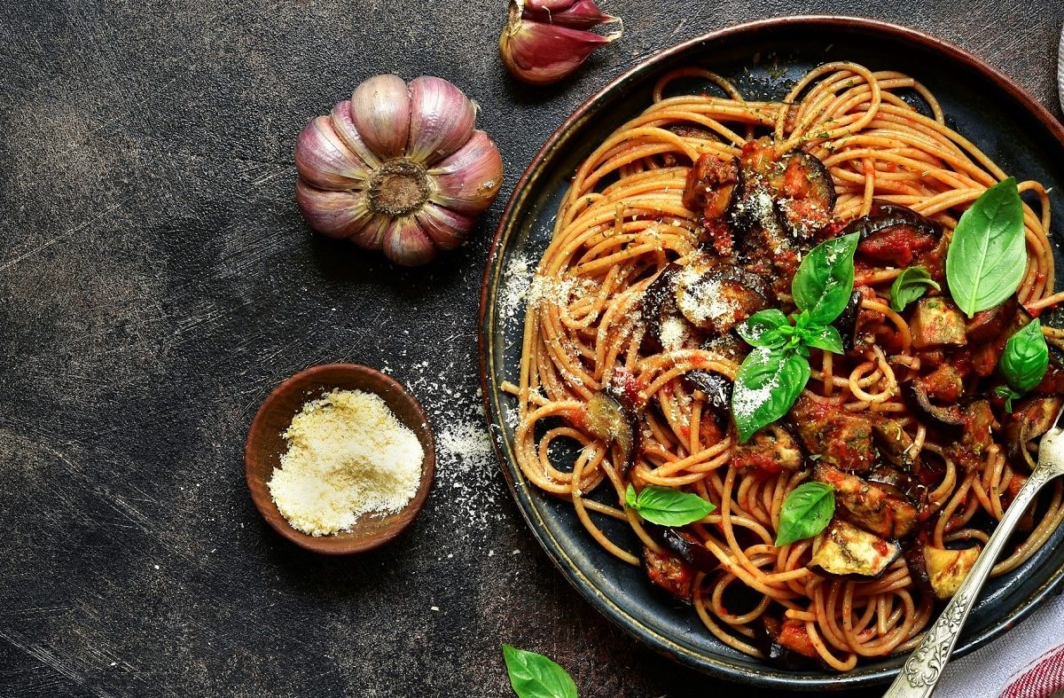 Spaghetti mit italienischer Norma-Sauce auf dunklem Teller. Das Ganze steht auf dunklem Untergrund und ist umgeben von lila Knoblauch und einem Schälchen Hartkäse.