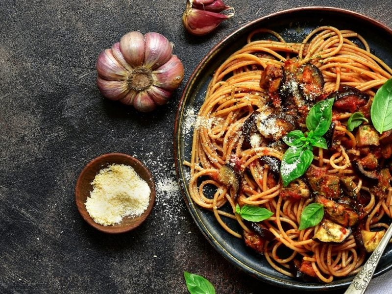 Spaghetti mit italienischer Norma-Sauce auf dunklem Teller. Das Ganze steht auf dunklem Untergrund und ist umgeben von lila Knoblauch und einem Schälchen Hartkäse.