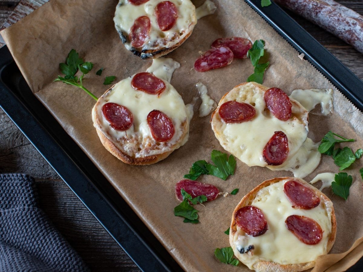 Pizza-Brötchen belegt mit Käse und Salami auf einem mit Backpapier ausgelegtem Blech.