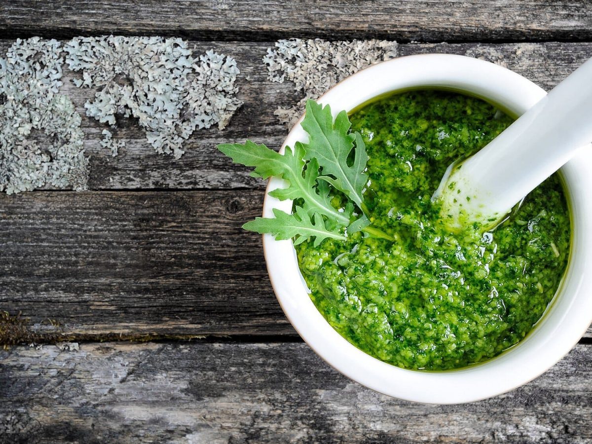 Rucola-Pesto in weißer Schale mit Mörser auf dunklem Holzuntergrund.