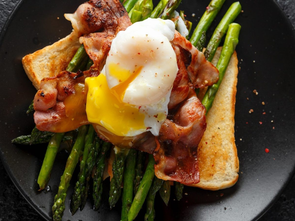 Eggs Benedict mit Spargel und Speck auf einem schwarzen Teller. Dieser steht auf dunklem untergrund.