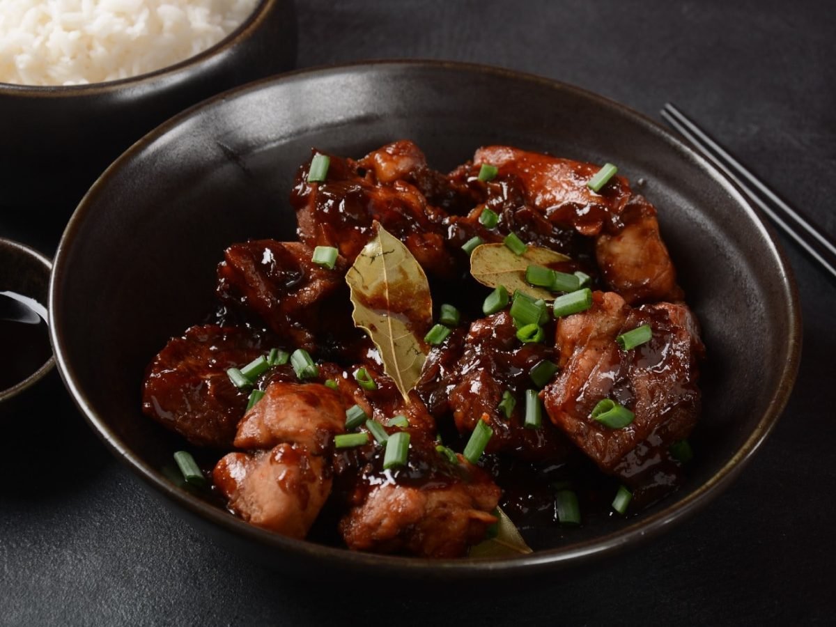 Eine schwarze Schale mit Adobo und Reis im Hintergrund auf schwarzem Tisch.