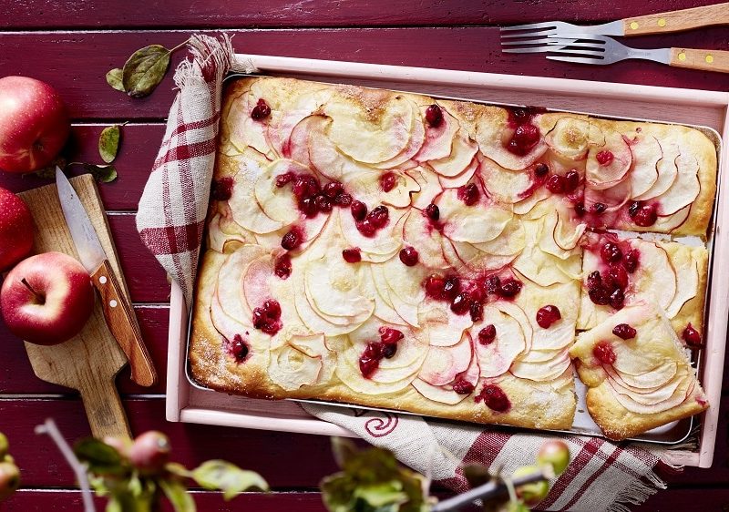 Aus der Vogelperspektive: Auf einem hölzernem Untergrund steht ein Apfel-Cranberry-Kuchen vom Blech. Daneben liegen drei Äpfel und Besteck..