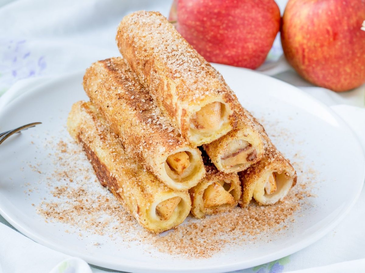 Apfel-Toast-Rollen auf einem weißen Teller. Daneben Besteck und verschiedene Zutaten.