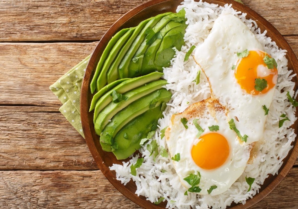 Arroz con huevo frito: Weißer Reis mit zwei Spiegeleiern und Avocado auf einem hölzernen Hintergrund