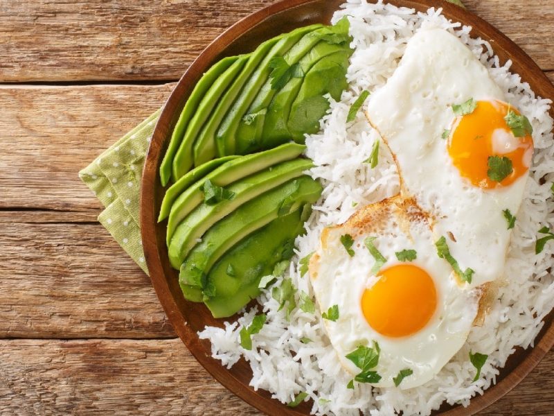 Arroz con huevo frito: Weißer Reis mit zwei Spiegeleiern und Avocado auf einem hölzernen Hintergrund