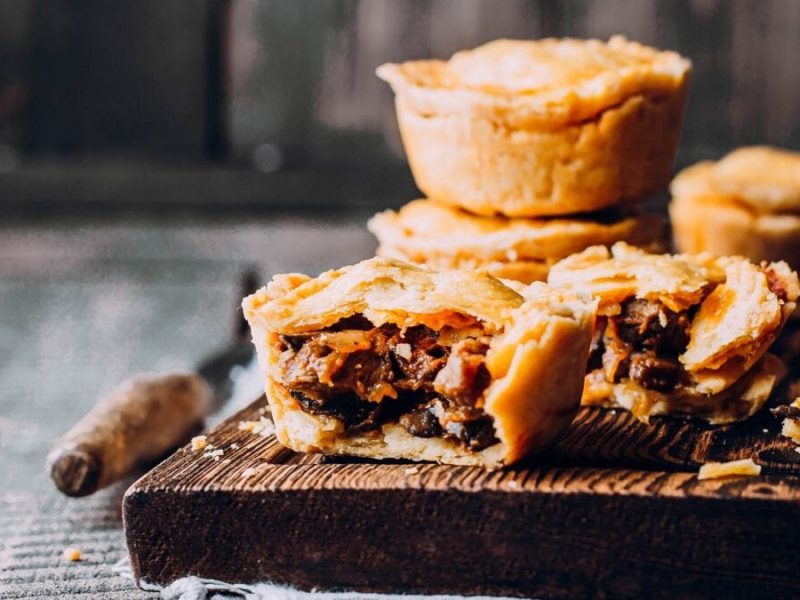 Australischer Meat Pie aus der Serie Bridgerton in angeschnitten, gestapelt und einfacher Form auf Holzbrett und grauem Untergrund. Daneben Messer mit Holzgriff. Frontalansicht.