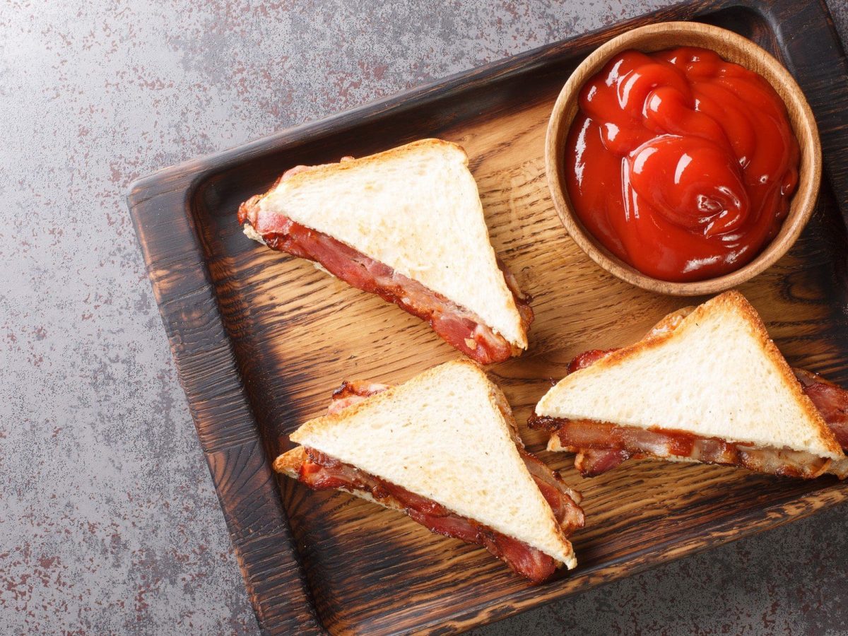 Bacon Butty Sandwich auf einem hölzernen Tablett. Daneben eine Schale mit Ketchup.