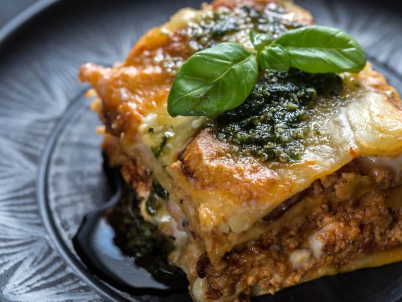 Bärlauch-Lasagne mit selbst gemachtem Bärlauchpesto auf grauem Teller mit Ornamenten auf grauem Untergrund. Basilikumblatt als Dekoration. Draufsicht.