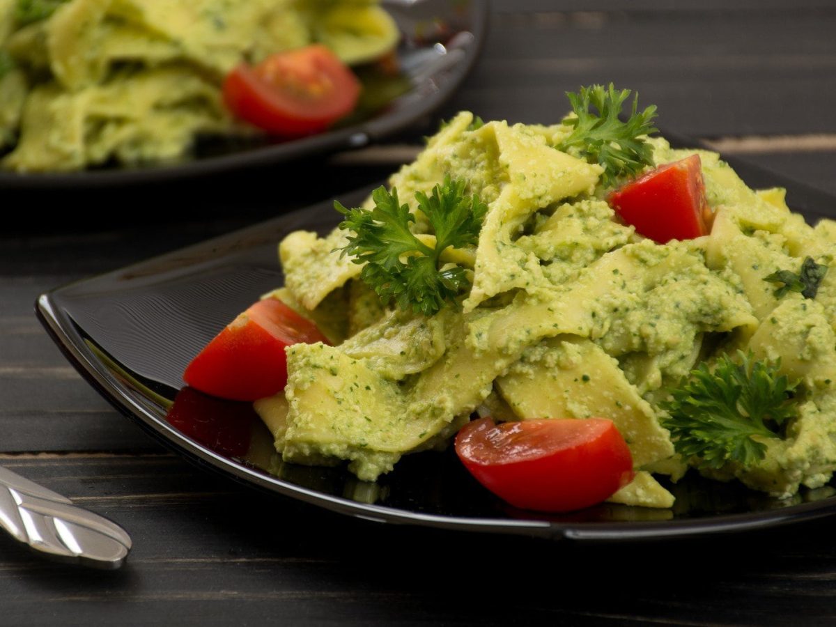 Zwei Teller Bandnudeln mit Cashew-Spinat-Sauce, Tomaten und Petersilie auf dunklem Holz mit Besteck.