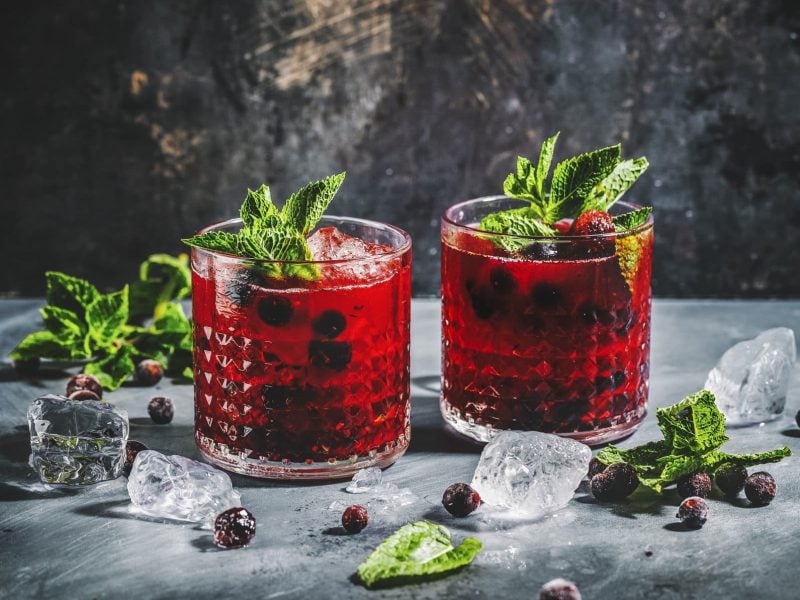 Zwei Gläser mit Blaubeer-Mojito und Minze garniert auf grauem Tisch mit Eiswürfeln.