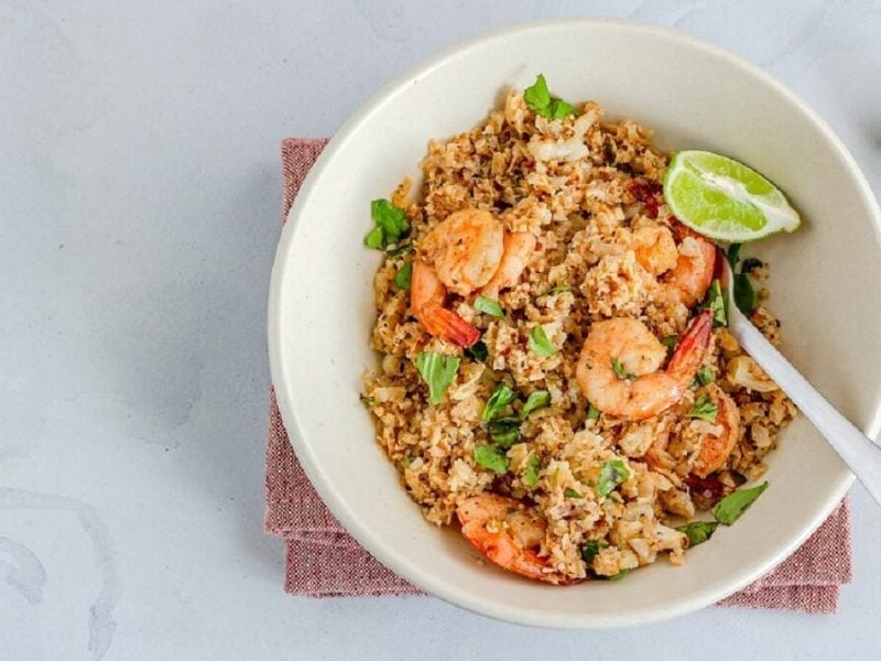 auf einem Tisch steht eine weiße Schüssel. In der Schüssel ist eine Mahlzeit, die Blumenkohl-Shrimps-Pfanne mit Basilikum heißt. Daneben liegen Limettenschalen und Basilikumblätter. In der Blumenkohl-Shrimps-Pfanne mit Basilikum steckt ein Löffel.