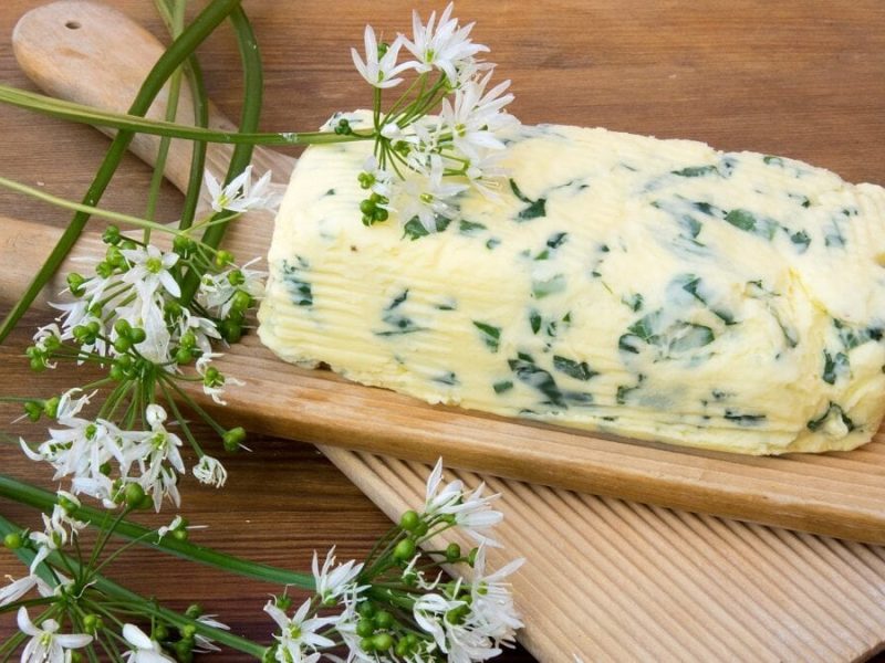 Auf einem Holzbrettchen liegt ein Stück Butter mit wildem Knoblauch. Daneben siist Bärlauch mit Blüten zu sehen.