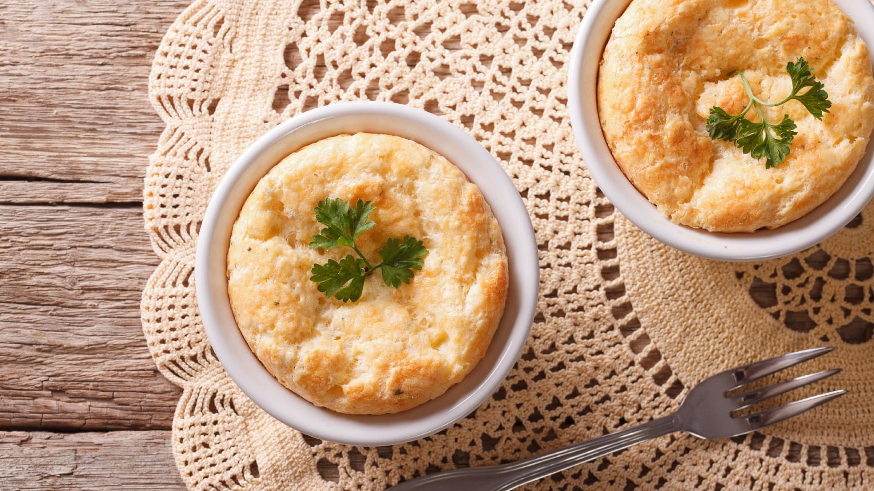 Zwei Cheddar-Souffles auf einem Holztisch mit beiger Tischdecke in Horizontalansicht von oben.