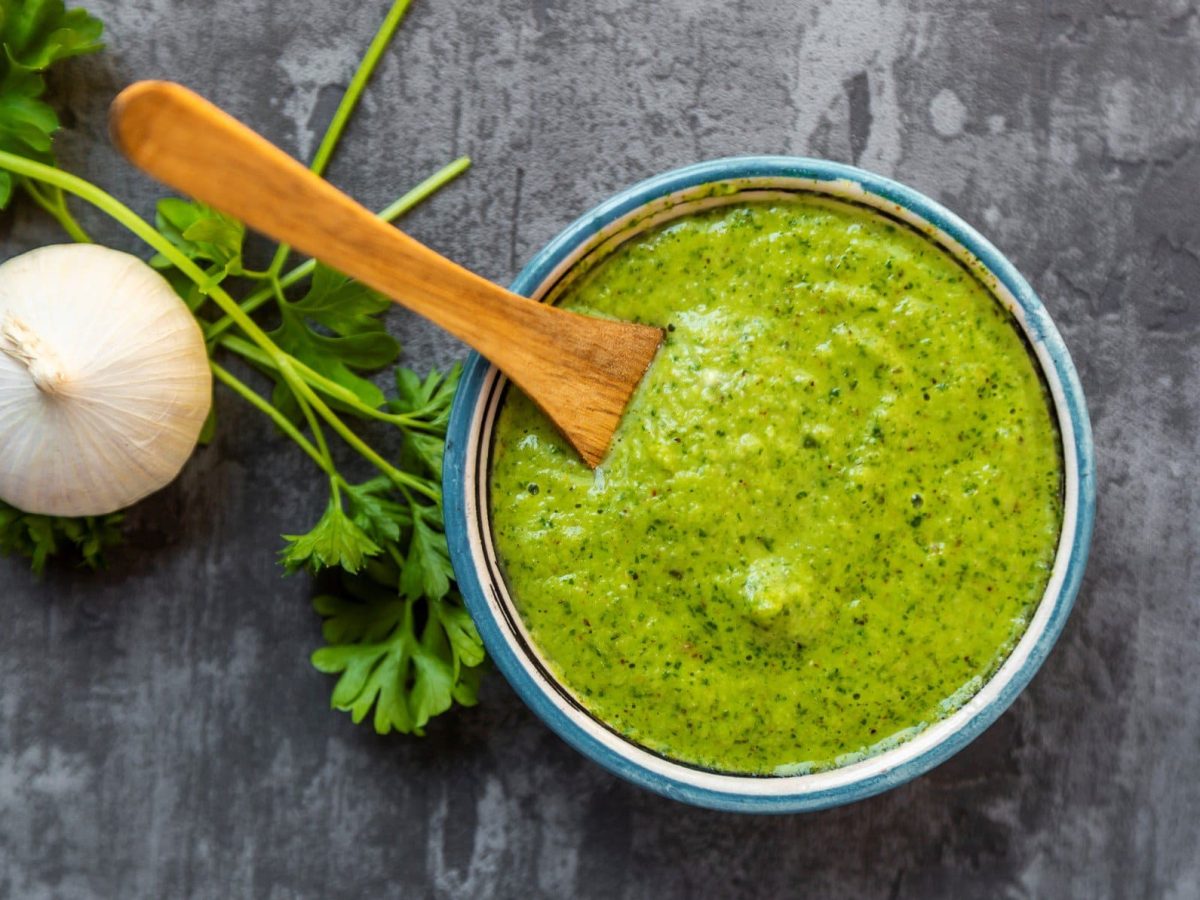 Eine hellblaue Schale mit Chimichurri auf grauem Untergrund. Links davon Petersilie und eine Knoblauchknolle.