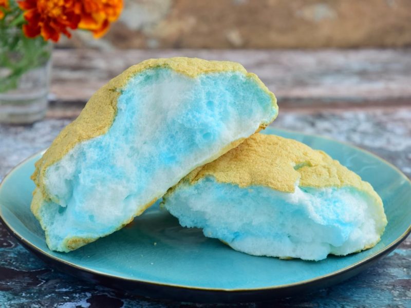 Ein blau-gelbes Brötchen auf einem Holztisch mit orangenen Blumen drauf.