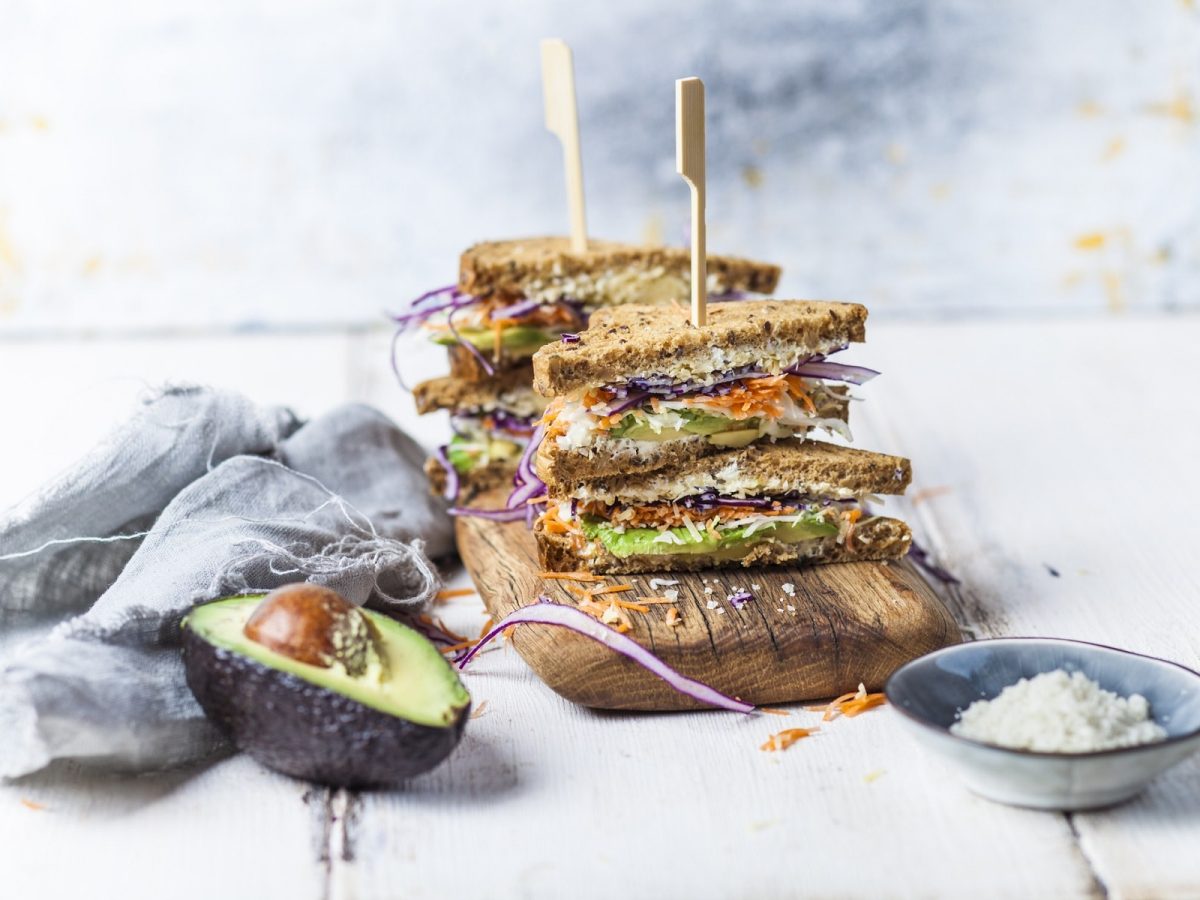 Zwei doppelte Sandwiches mit Holzstäbchen fixiert auf einem Brett, daneben liegen eine halbe Avocado und ein Geschirrtuch.