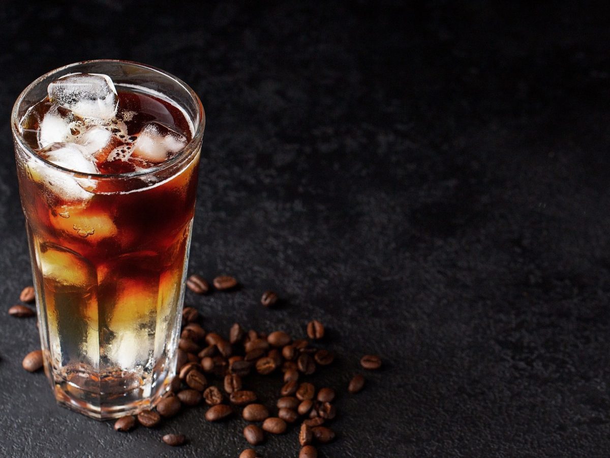 Ein Glas Espresso Tonic mit Eiswürfeln in dunklem Setting, daneben liegen Kaffeebohnen.