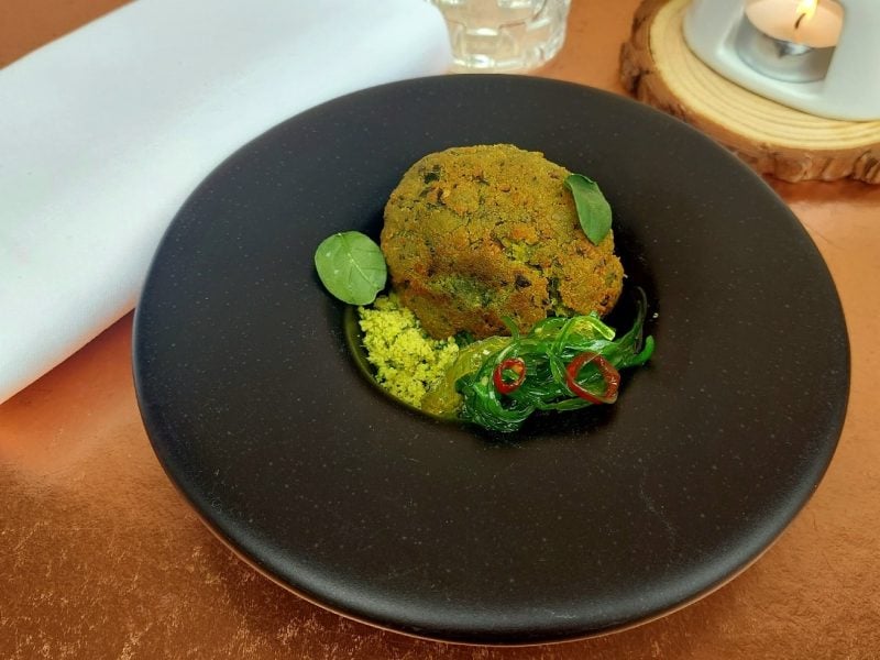 Grün paniertes Falafel-Ei mit Algensalat in einem schwarzen Teller auf einem braunen Tisch.