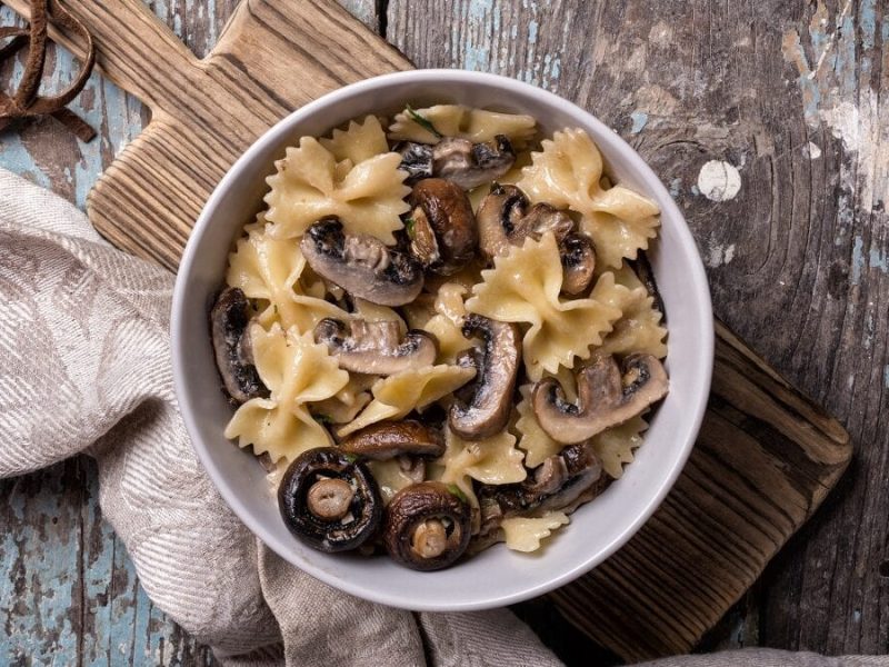 auf einem hölzernen Untergrund liegen ein Brettchen und ein Handtuch. Auf dem Brettchen steht eine große Schüssel mit Farfalle mit Champignons