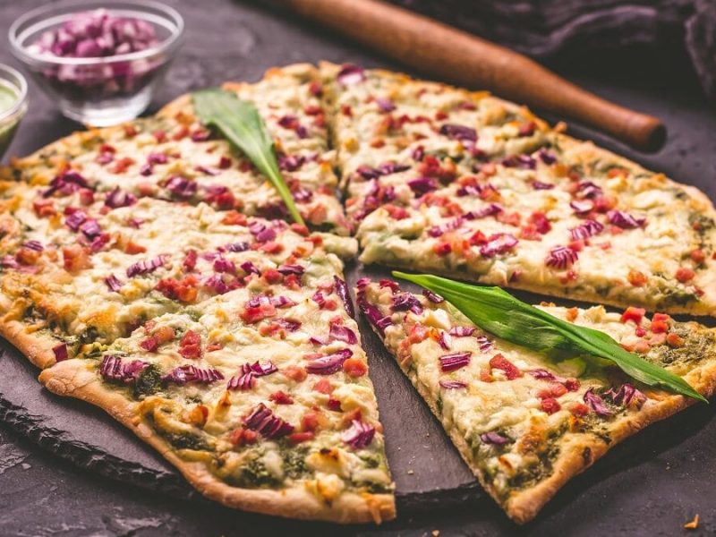 Auf einem Untergrund aus Schiefer liegt ein Flammkuchen mit Bärlauch. Daneben stehen zwei kleine Glasschüsseln, In er einer sind rote Zwiebeln und in der anderem eine grüne Creme.