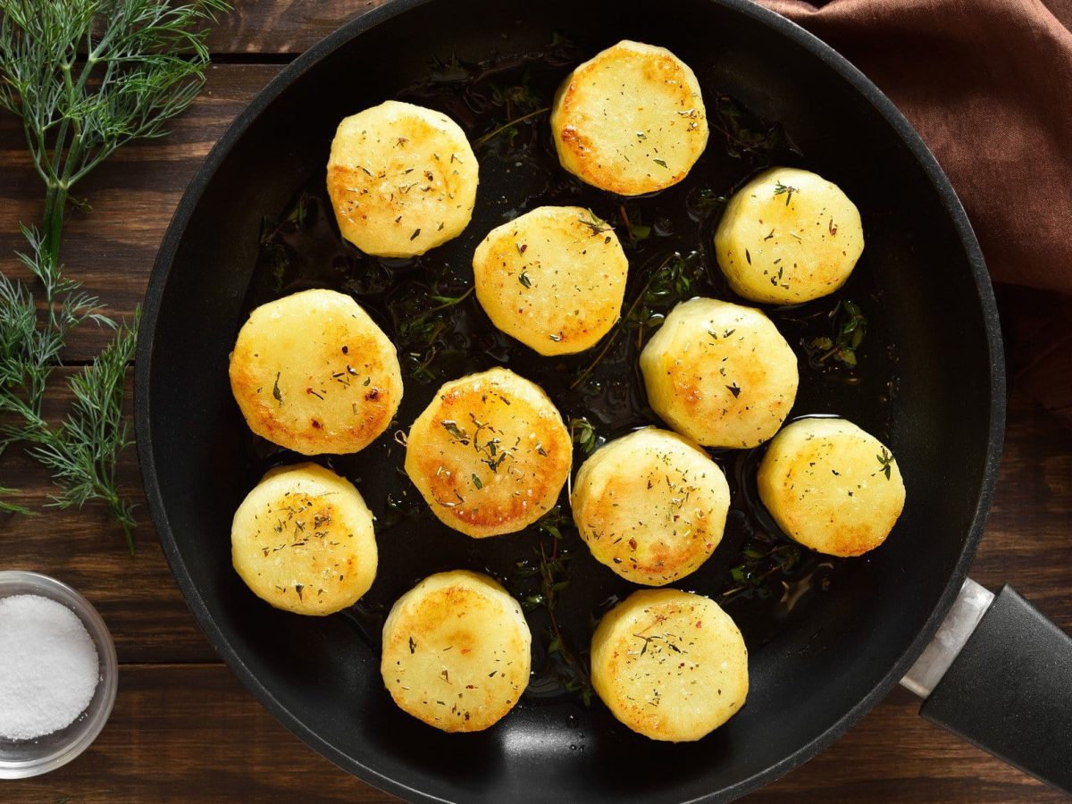 Fondant-Kartoffeln in einer Pfanne. Daneben liegt Rosmarin, eine Schale mit Salz und ein Küchentuch.