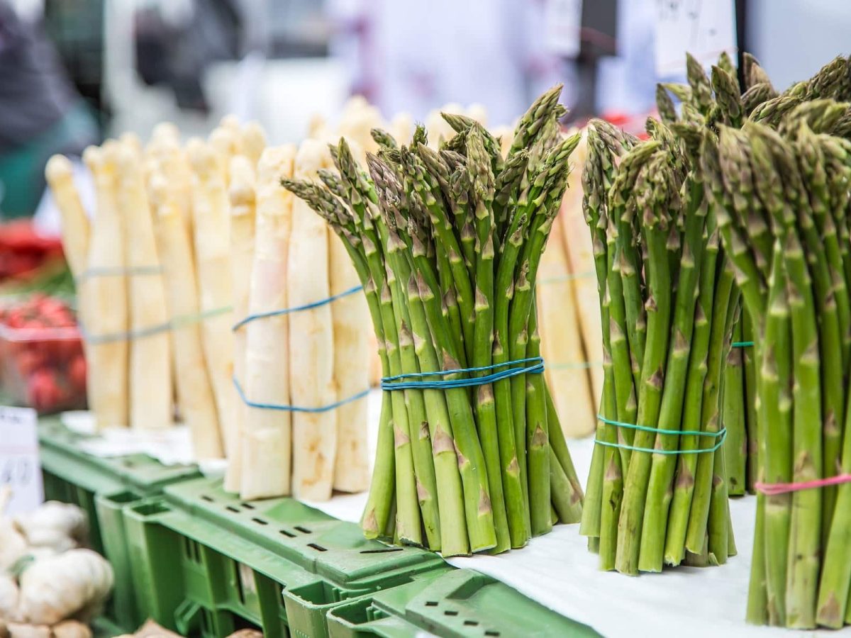 Grüner und weißer Spargel jeweils als Bund an einem Marktstand. Im Hintergrund sind Erdbeeren.