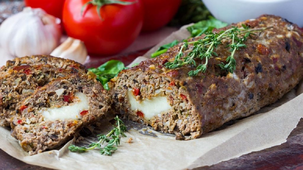 Gefüllter Hackbraten mit Käse und Schmorgemüse zum Muttertag auf weißem Teller mit frischem Thymian. Im Hintergrund Tomaten und Knoblauch. Frontalansicht.