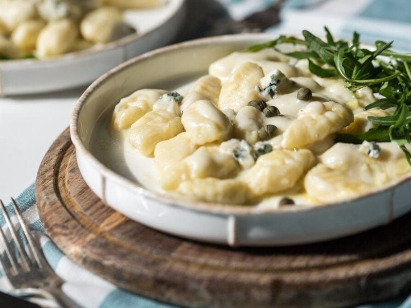 Auf einem Brettchen steht ein Teller mit Gnocchi mit Gorgonzola-Kapern-Sauce. Daneben lieen ein weiterer Teller mit der Pasta sowie eine Gabel.