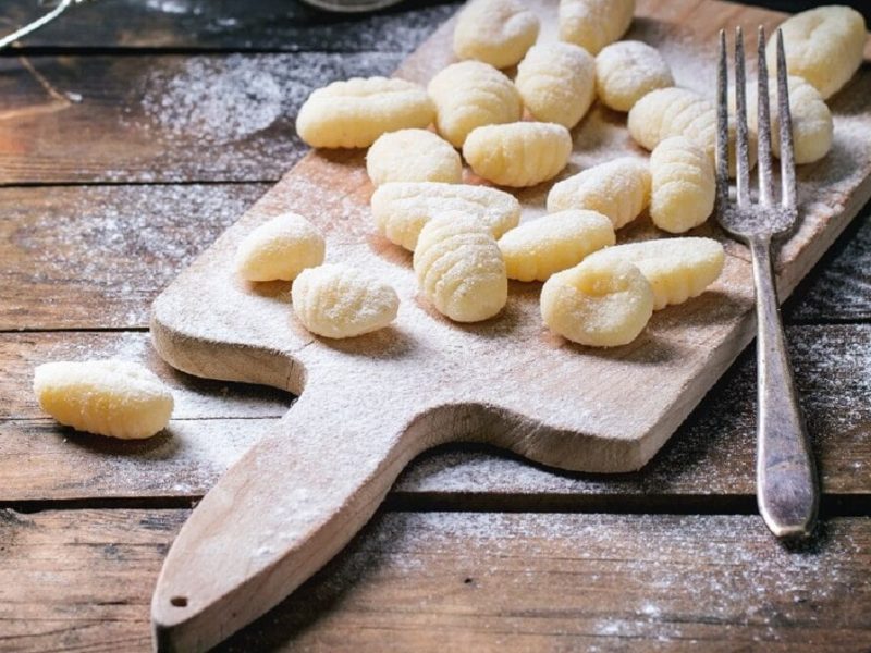 Auf einem holzbrett liegen selbst gemachte gnocchi, die mit Mehl bestäubt sind. daneben liegt eine Gabel.