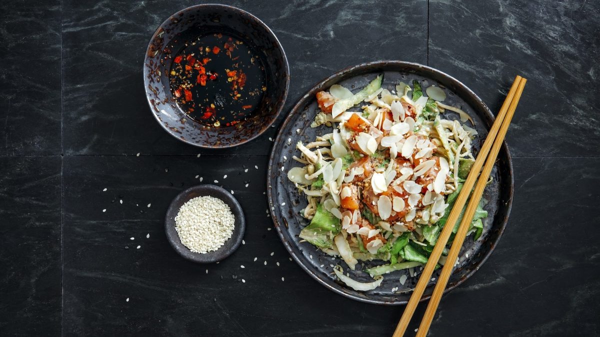 Japanischer Salat mit Lachs, Mandeln, Sesam, Ingwer, Avocado und Chili in einer Schale mit Stäbchen, dazu Chili-Öl und Sesamflocken auf einem steinernen Untergrund, von oben fotografiert.