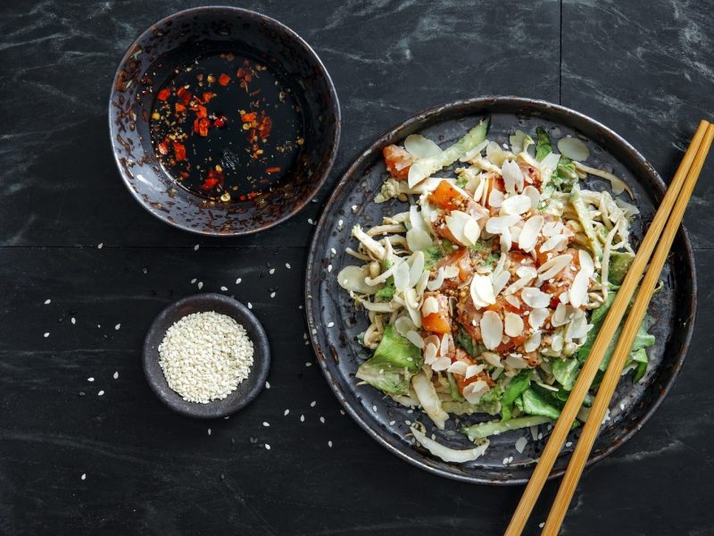 Japanischer Salat mit Lachs, Mandeln, Sesam, Ingwer, Avocado und Chili in einer Schale mit Stäbchen, dazu Chili-Öl und Sesamflocken auf einem steinernen Untergrund, von oben fotografiert.