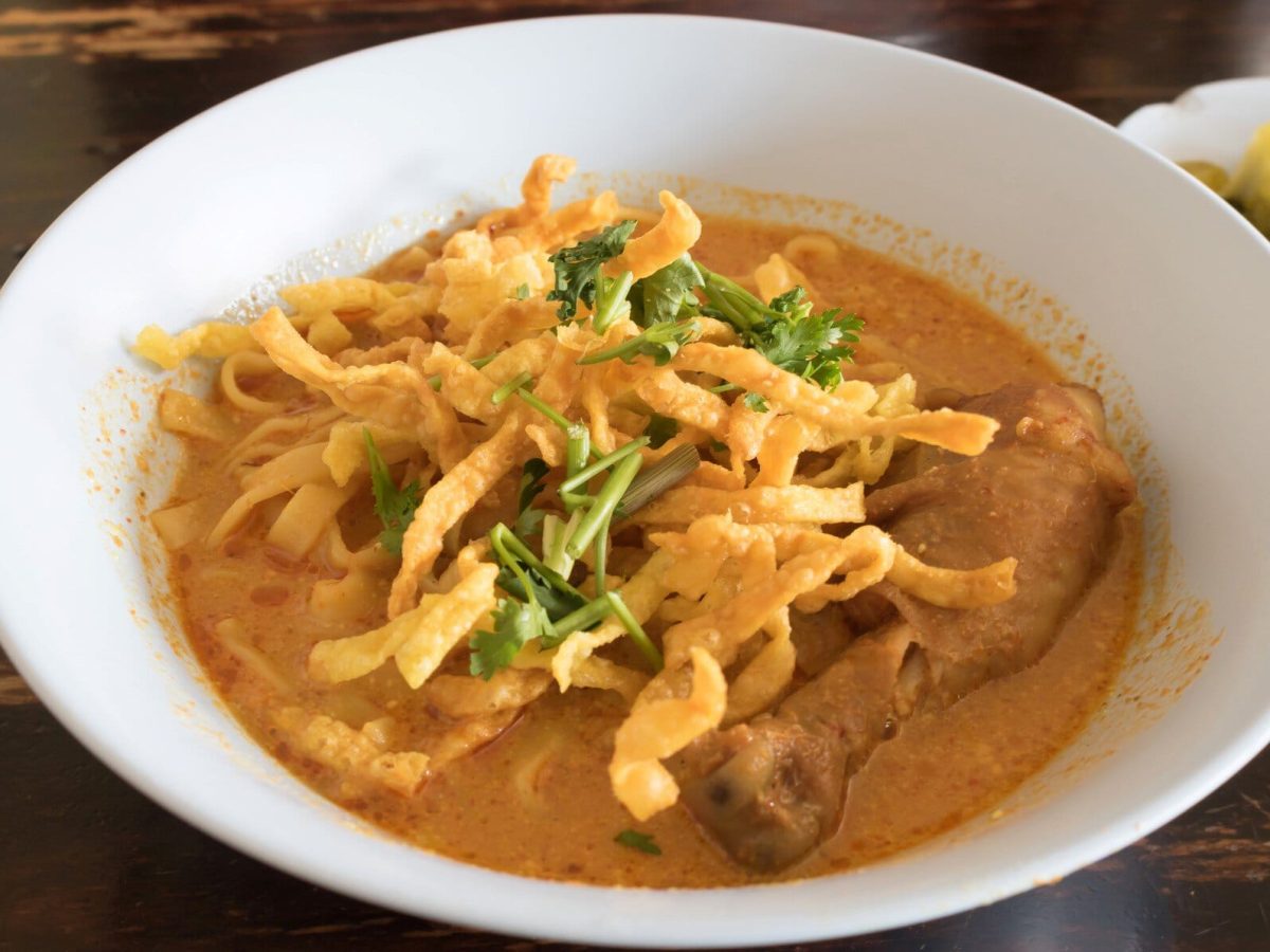 Eine weiße Schale Khao Soi in der Draufsicht auf einem Holztisch.