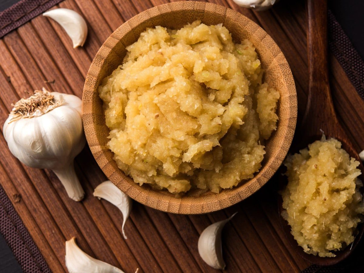 Geröstete Knoblauchpaste auf Holzbrett auf dunklem Untergrund. Daneben Holzlöffel mit der Paste und Knoblauchzehen verteilt. Aufnahme aus der Vogelperspektive.