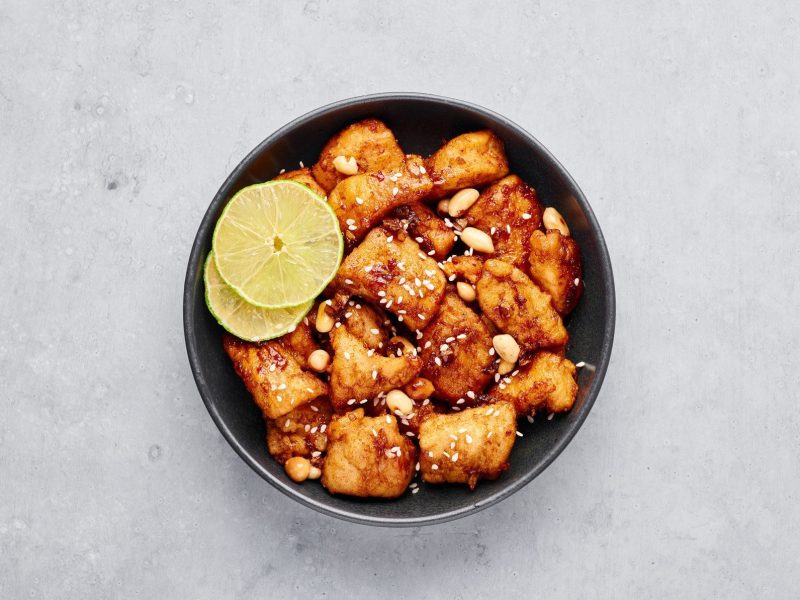 Koranisches Dakgangjeong Korean Fried Chicken in einer schwarzen Schale mit Sesam, Erdnüssen und Zitronenscheiben garniert auf grauem Untergrund. Aufnahme aus der Vogelperspektive.