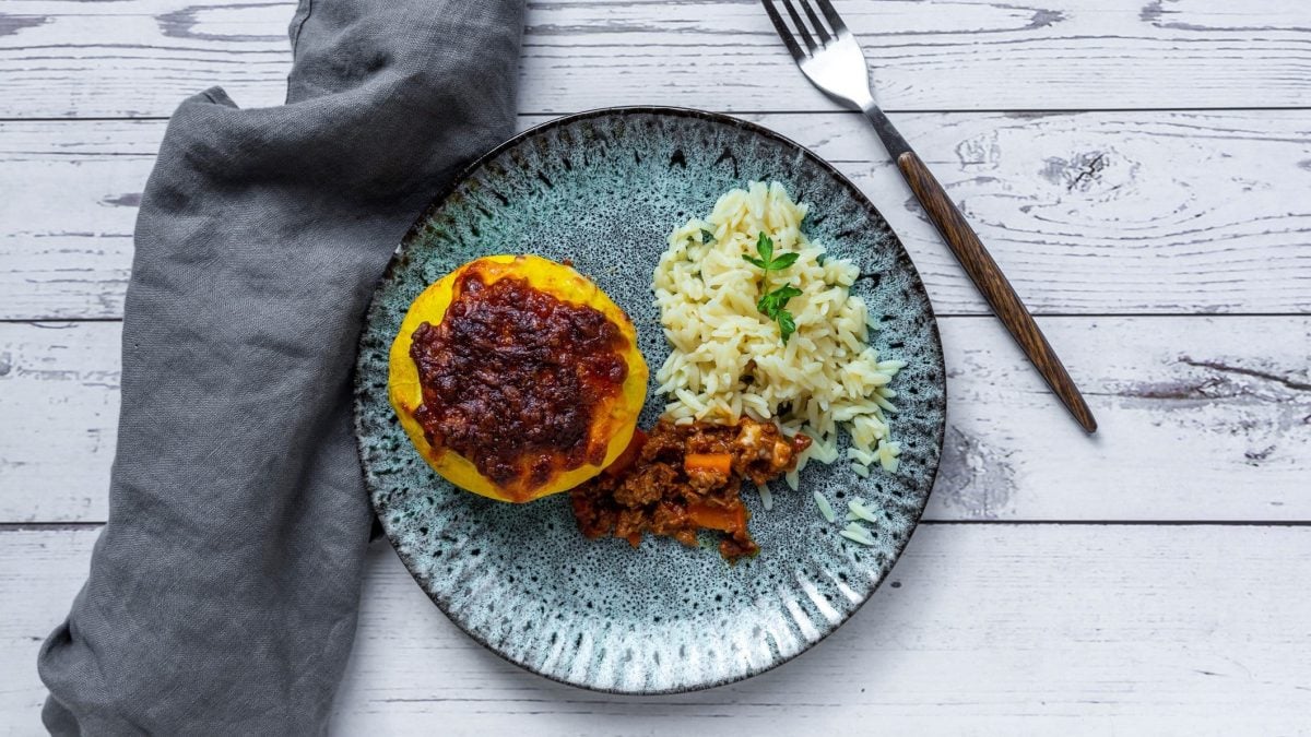 Ein ausgehöhlter Kürbis mit veganer Bolognese gefüllt auf einem blauen Teller mit Reis an der rechten Seite. Alles auf einem weißen Holztisch mit grauer Decke.