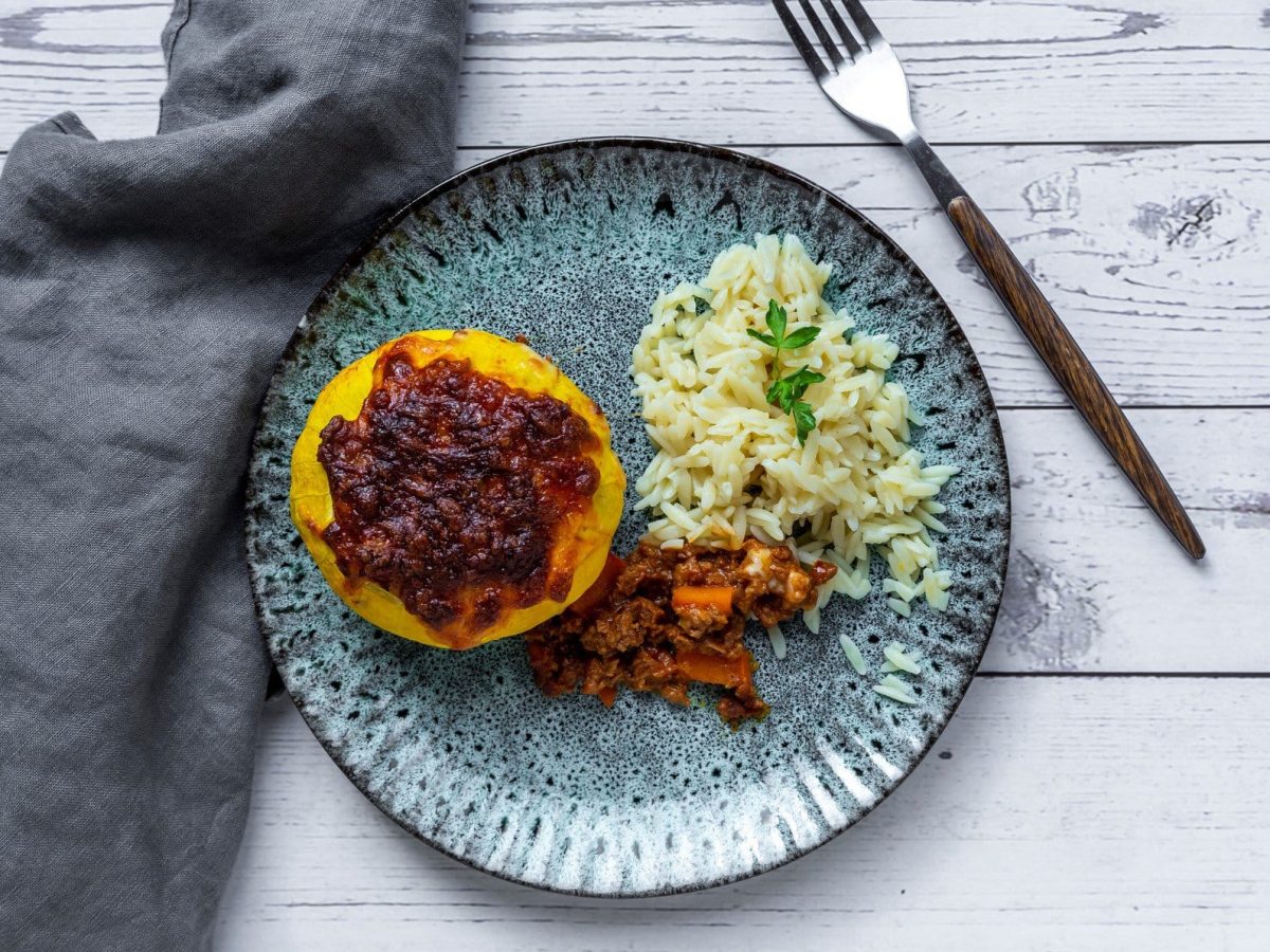 Ein ausgehöhlter Kürbis mit veganer Bolognese gefüllt auf einem blauen Teller mit Reis an der rechten Seite. Alles auf einem weißen Holztisch mit grauer Decke.