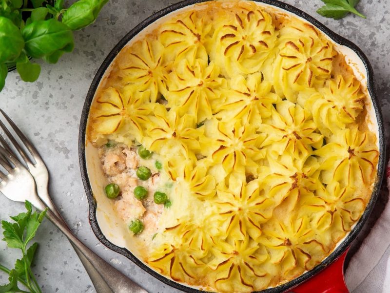 Lachs-Pie mit Erbsen und Kartoffelpüree überbacken, serviert in einer runden Auflaufform, serviert auf einem hellen Tisch.