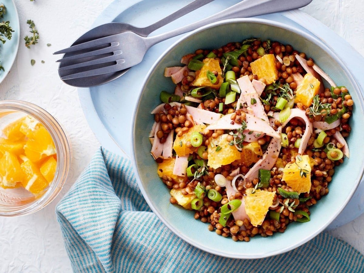 Linsensalat mit Fruchtnote, garniert mit frischem Thymian, feiner Vinaigrette und Kasseler-Streifen in einer hellen Schale auf weißem Untergrund