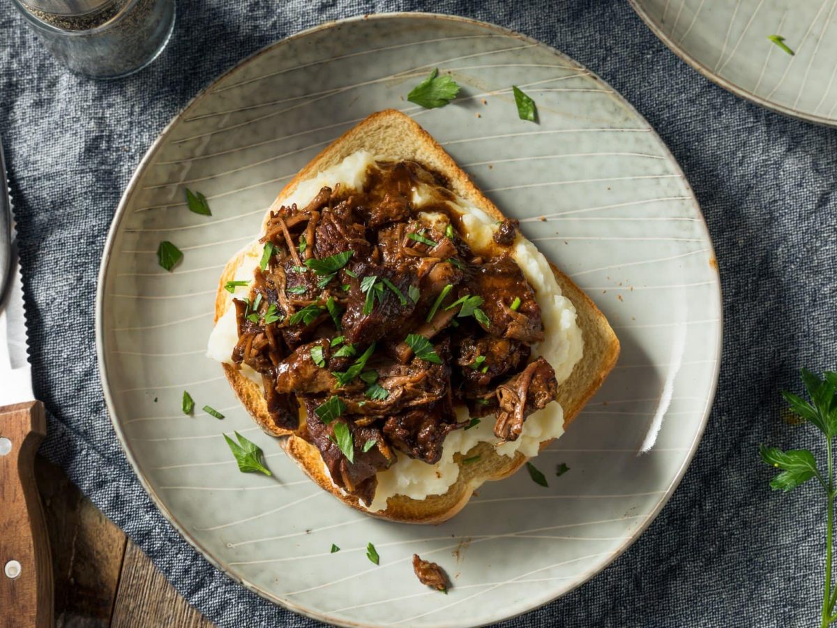 Manhattan Roast Sandwich auf hellem Teller mit Petersilie bestreut. Daneben Messer und Gabel. Rechts im Bild auf grauem Untergrund Petersilie. Vogelperspektive.