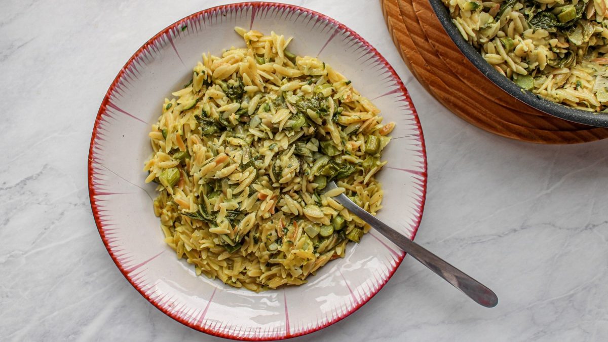 Ein Teller Orzopasta mit grünem Spargel und Spinat in der Draufsicht, daneben steht der Topf mit noch mehr Pasta.