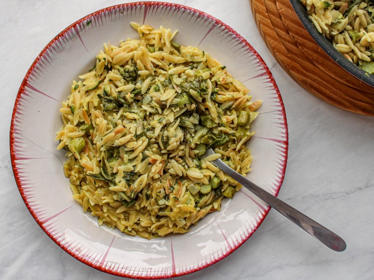 Ein Teller Orzopasta mit grünem Spargel und Spinat in der Draufsicht, daneben steht der Topf mit noch mehr Pasta.