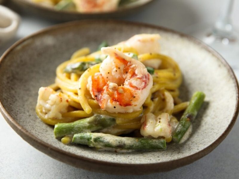 Pasta mit grünem Spargel und Gamberi in Limettensauce auf grauem Pastateller und hellem Tisch. Im Hintergrund angedeutet Weinglas und ein weiterer Teller. Draufsicht.