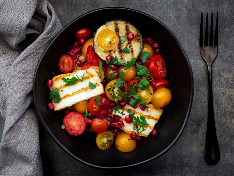 Eine schwarze Schale mit dem Tomatensalat und gegrillter Aubergine und Halloumi auf grauem Untergrund. Daneben ein graues Tuch und eine schwarze Gabel.