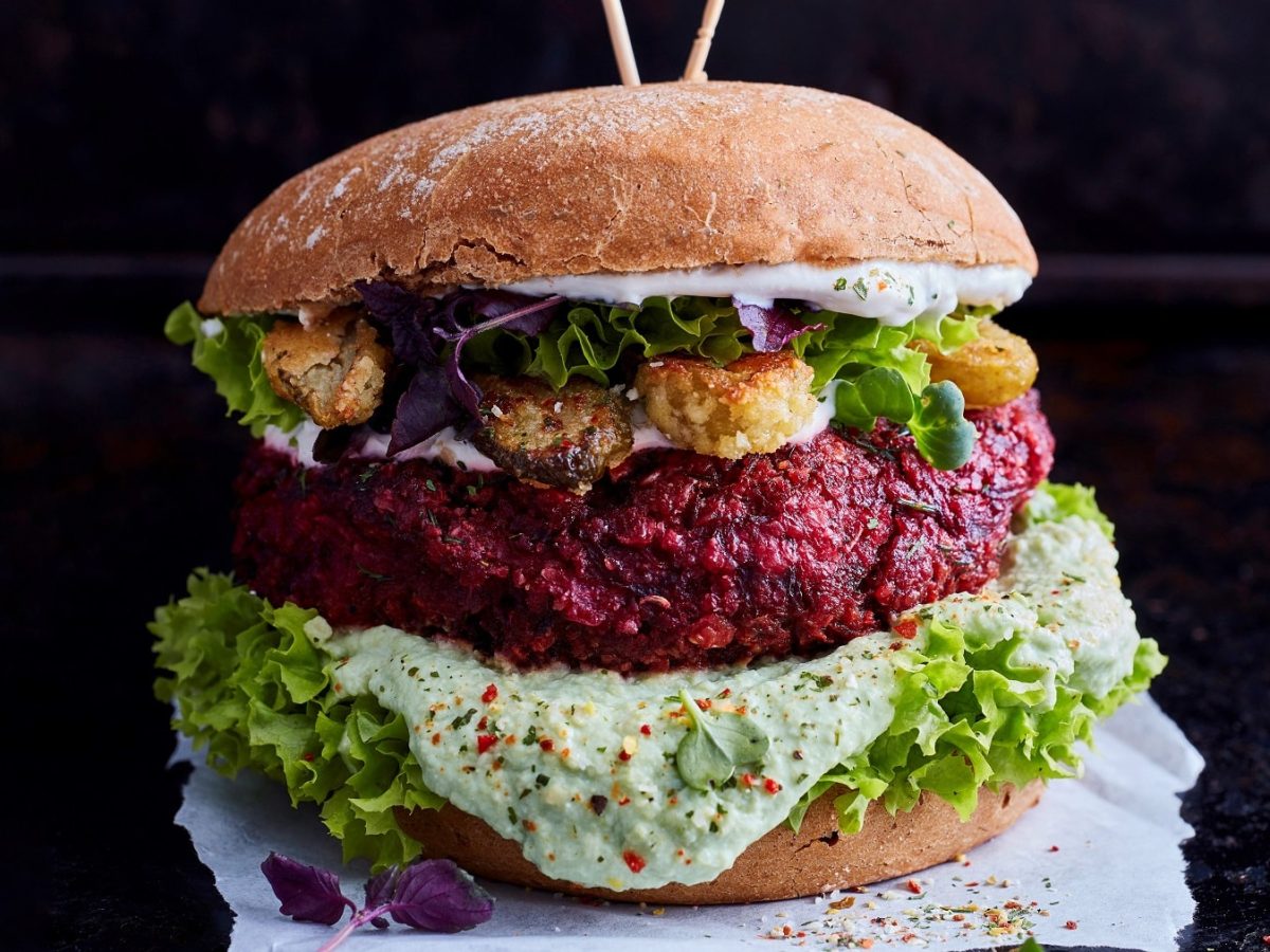 Ein Burger mit Namen "Purple Bliss Burger" mit rotem Bratling, Salat, weißer Kräutersauce und frittierten Gurken.